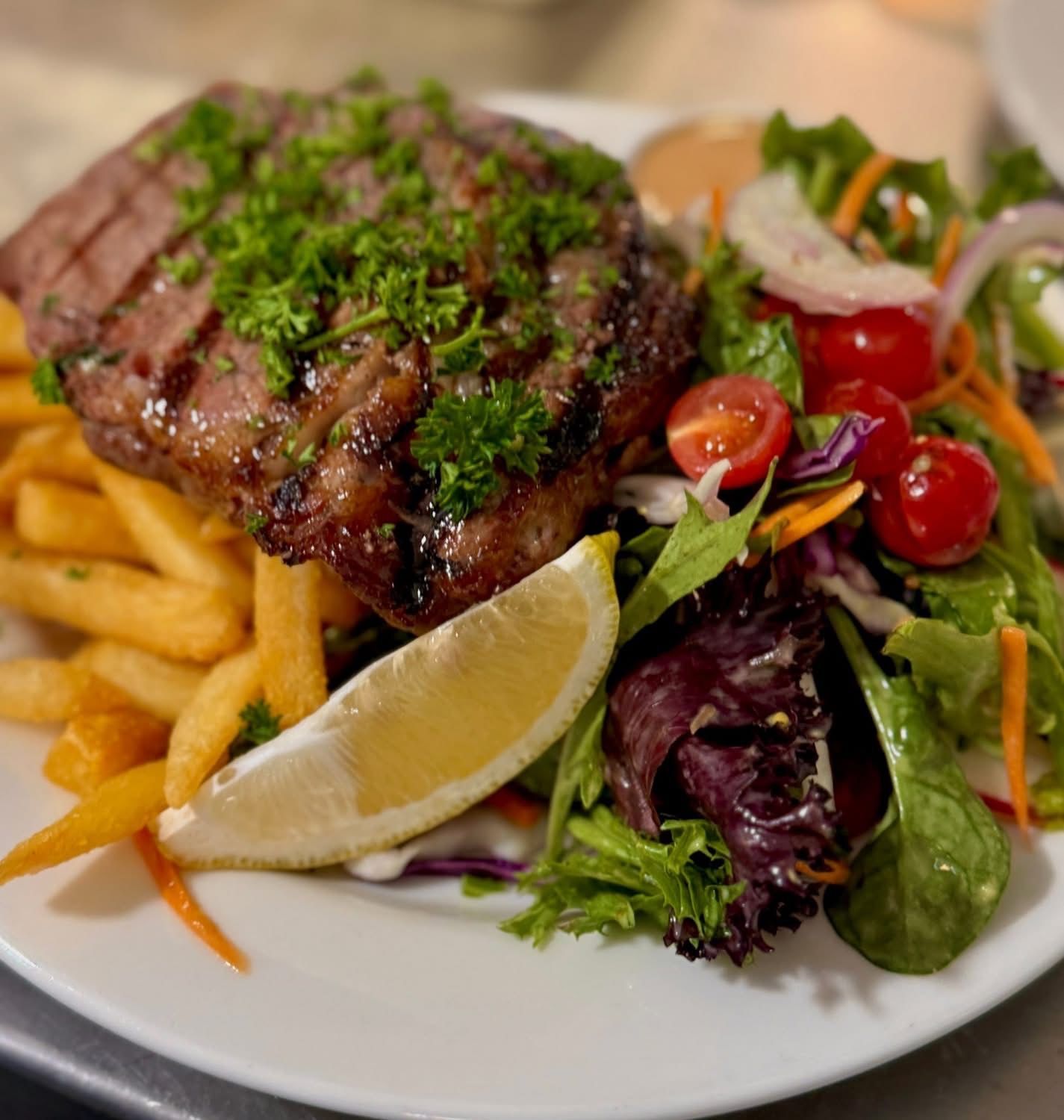 Grilled steak with fries, salad, and lemon wedge on a white plate.