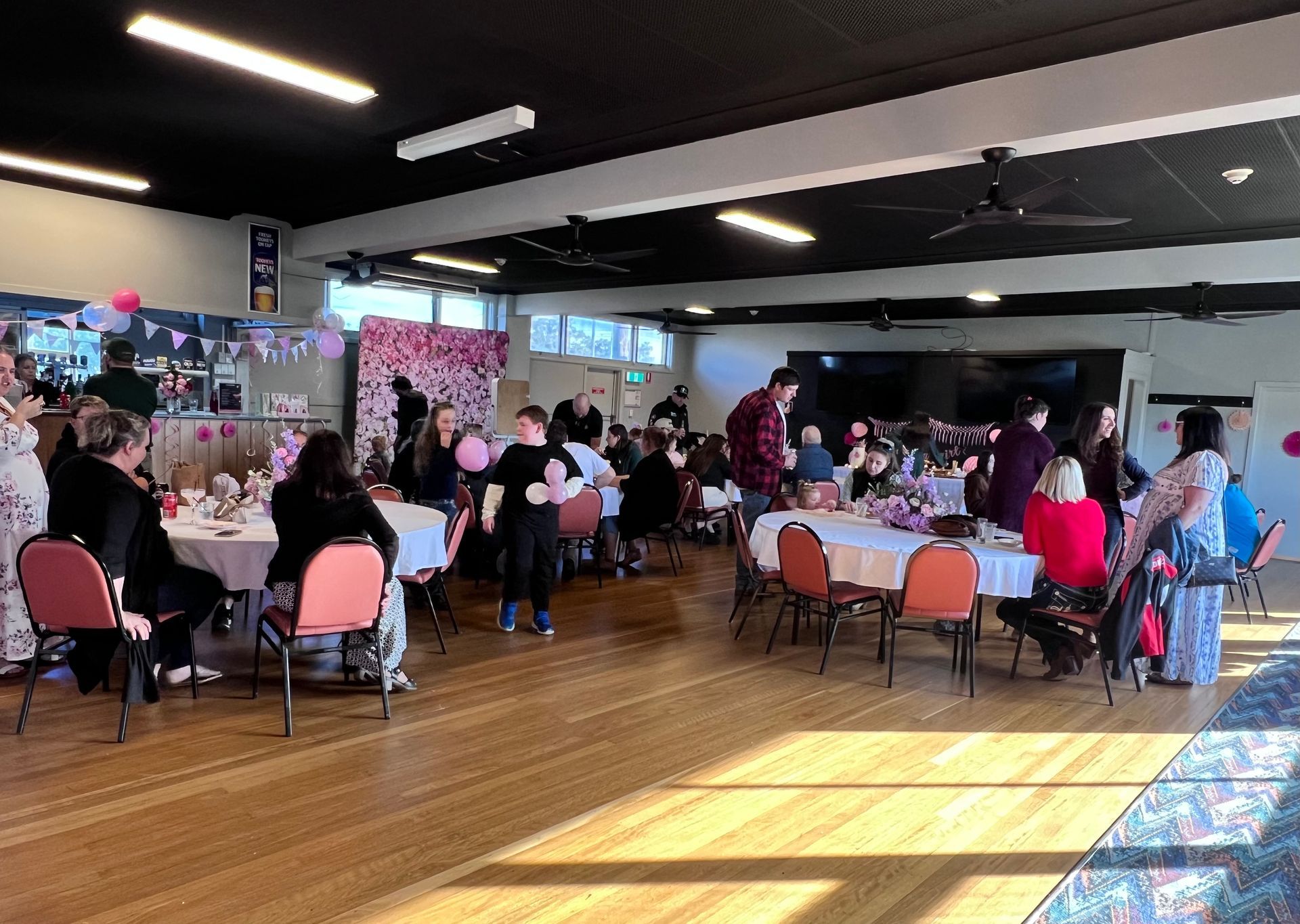 Event hall with tables, chairs, and people; pink floral backdrop, sunlight.