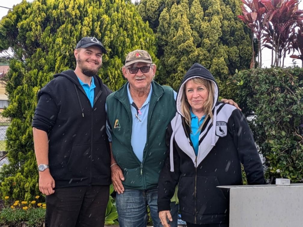 Three people standing outdoors. Man in cap, middle-aged man, woman in hooded jacket. Overcast, green shrubbery.