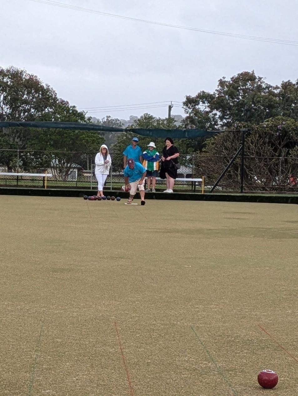 People playing lawn bowls on a green field under a cloudy sky. One person bowls. Others watch.