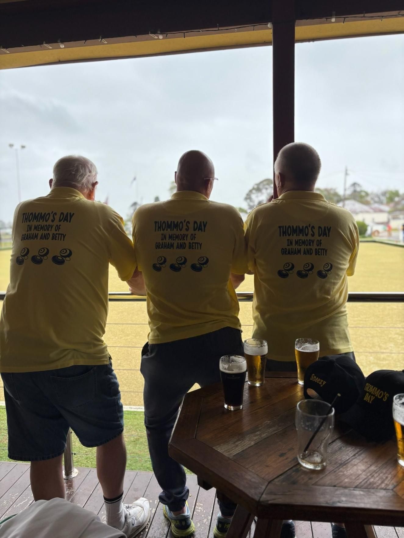 Three men in yellow shirts with text, standing at a bar looking out at a lawn bowls green.