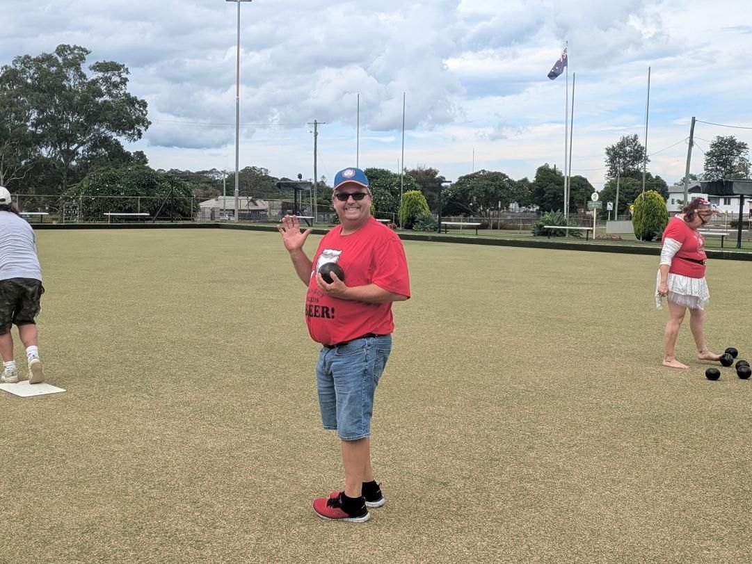 Person waving while holding a bowling ball on a grassy bowling green. Two other people are visible.