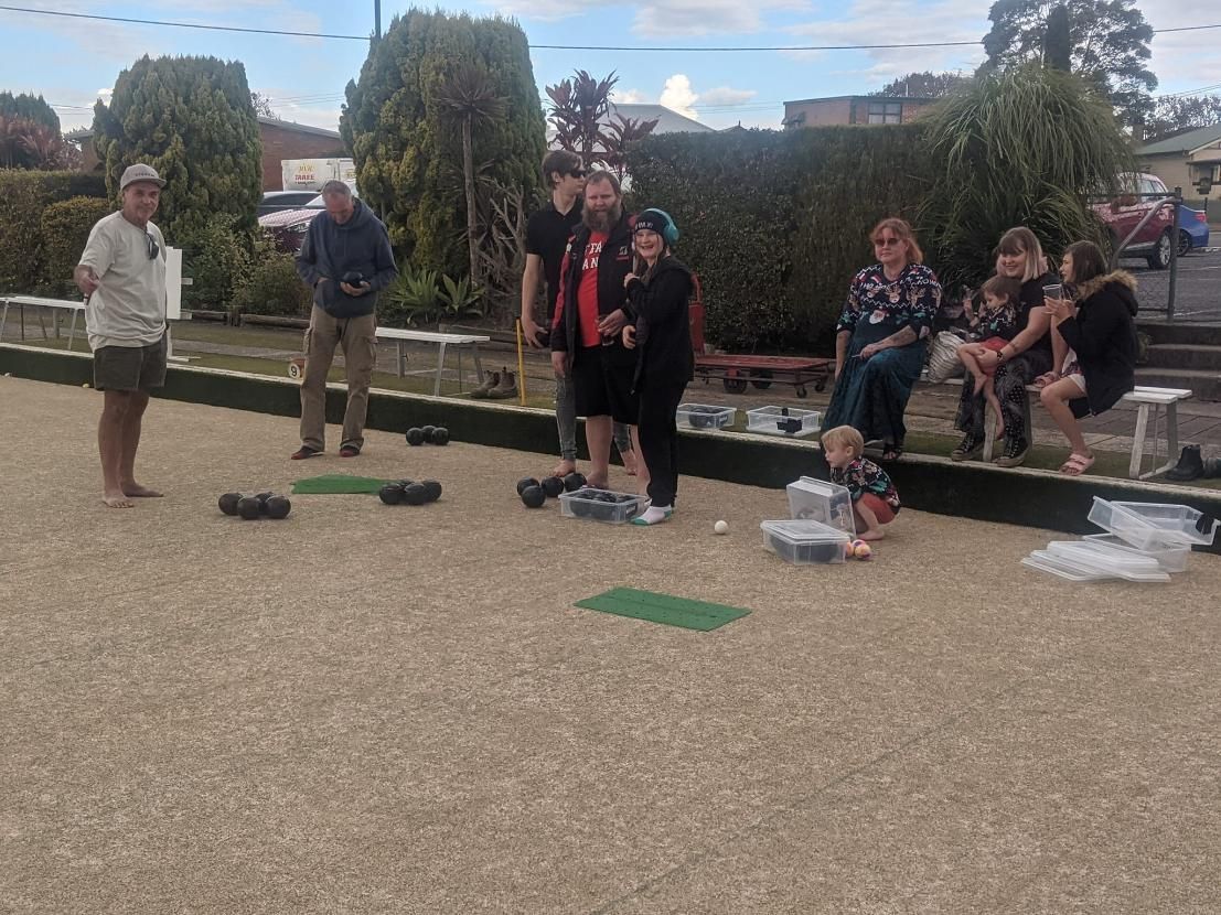 People playing lawn bowls outdoors; green and beige tones. Some are seated, others standing and observing.