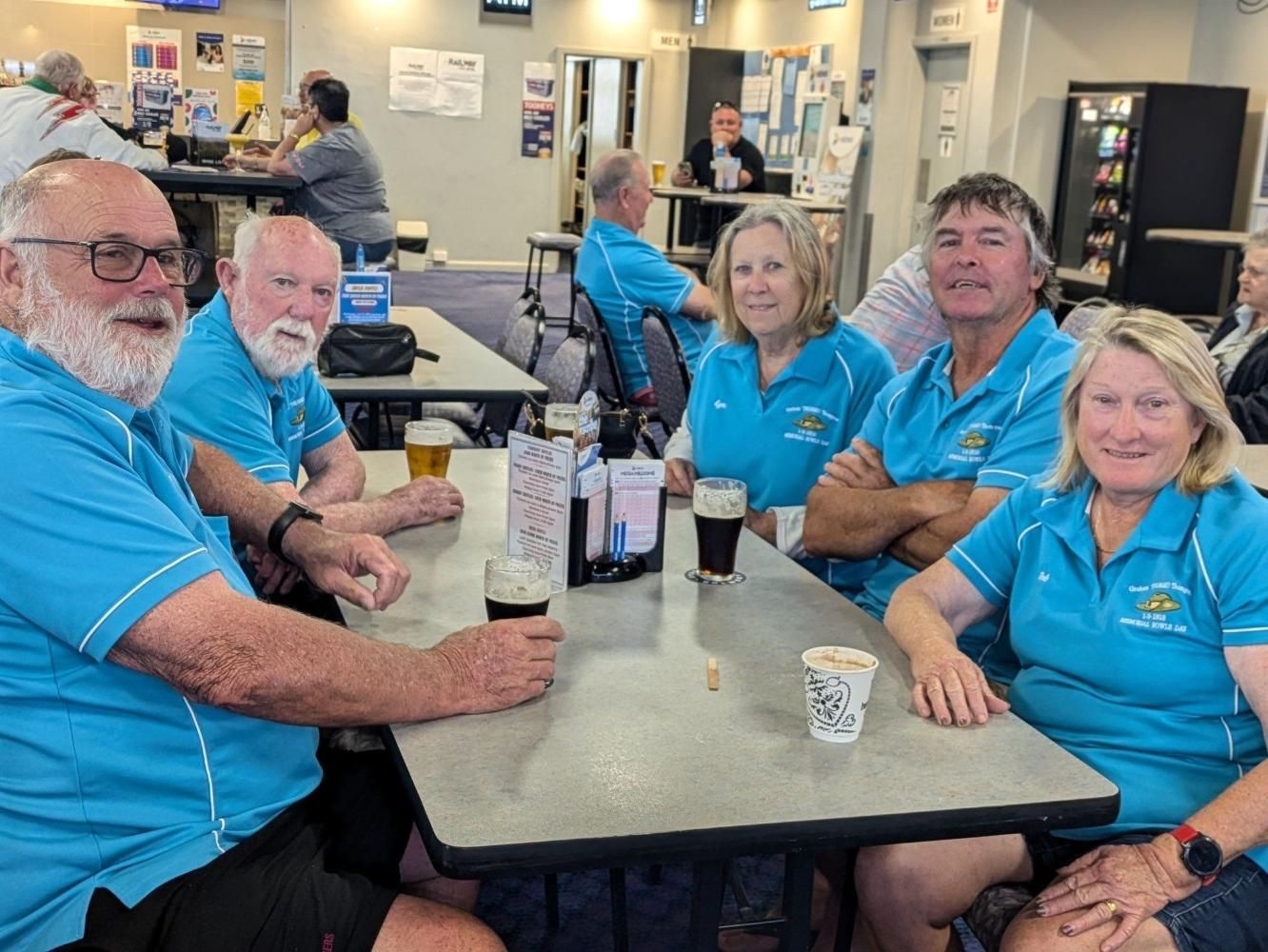 Group of people in blue shirts at a table, some with drinks, in a casual setting.
