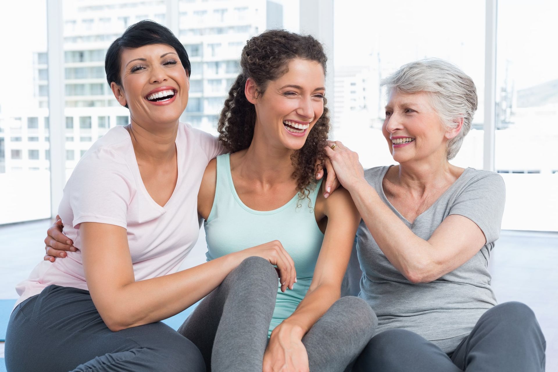 Three women in activewear laugh together. Two have arms around each other. Bright, spacious setting.