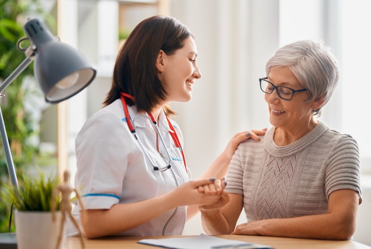A smiling healthcare provider comforts an elderly patient. They are holding hands and sitting at a desk.