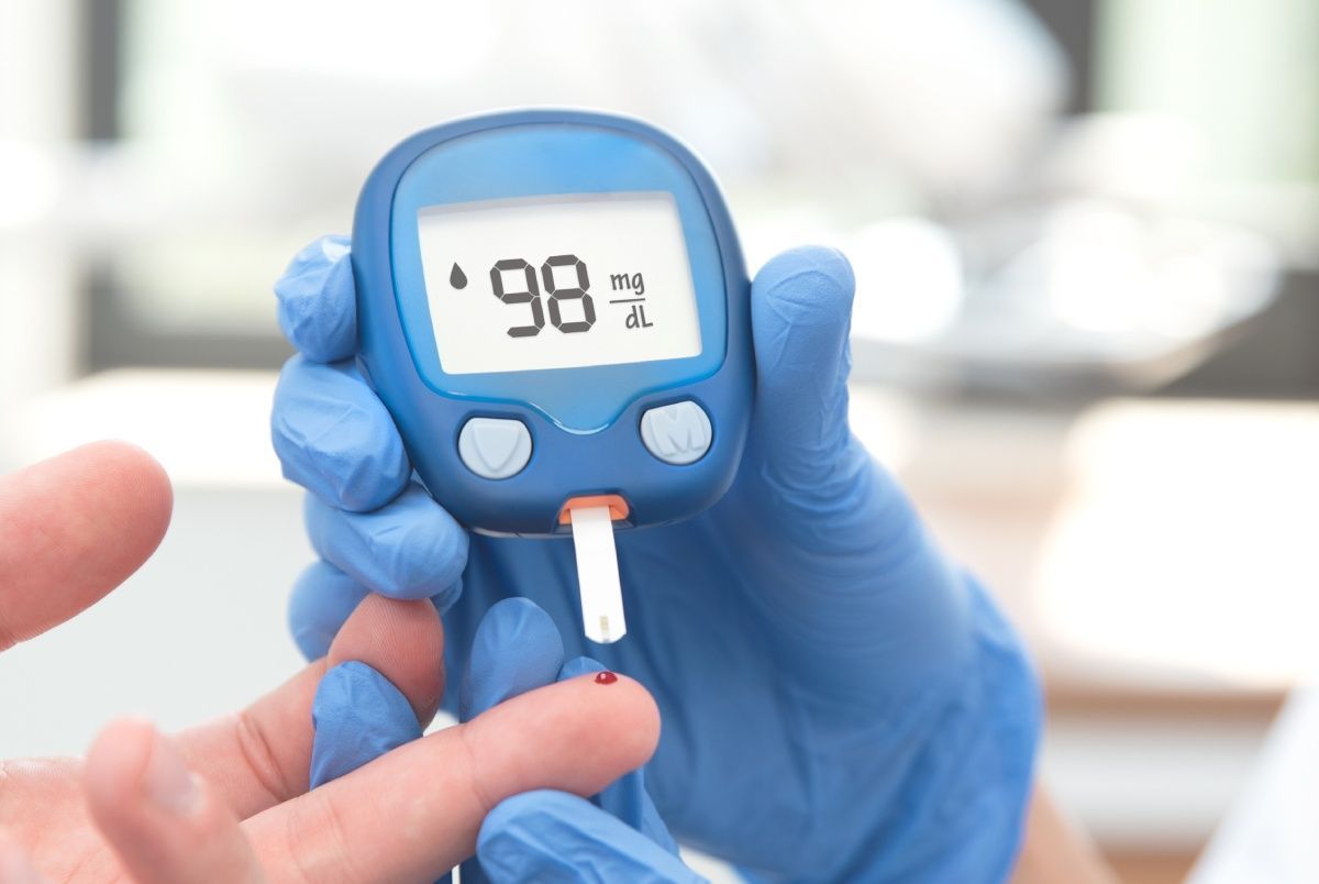 Person's finger pricked, being tested for blood glucose level using a glucometer; reading shows 98 mg/dL.