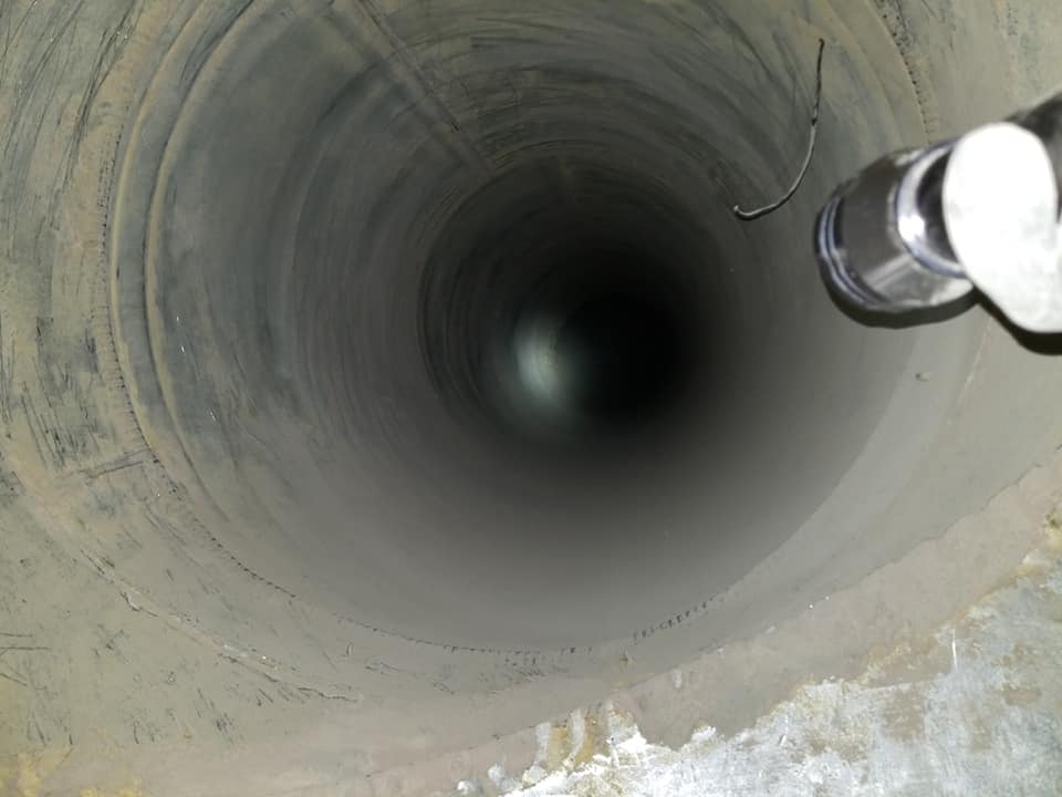 Vue plongeante sur un puits ou un conduit cylindrique en béton profond, éclairé par une lampe torche en haut à droite.