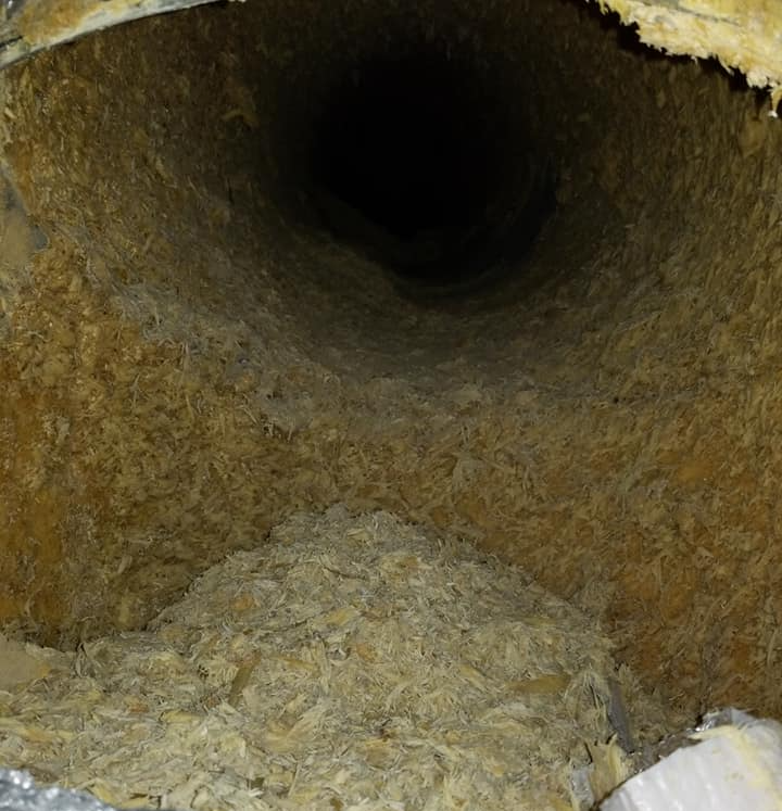 Vue intérieure d'un conduit de ventilation rond et poussiéreux, dont l'intérieur est tapissé d'isolant brun et au fond duquel s'entassent des débris.