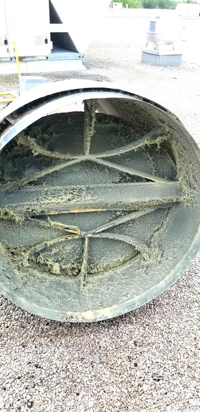 Un conduit d'échappement circulaire en métal, dont la grille est obstruée par des débris, est vu à l'extérieur sur une surface de gravier.