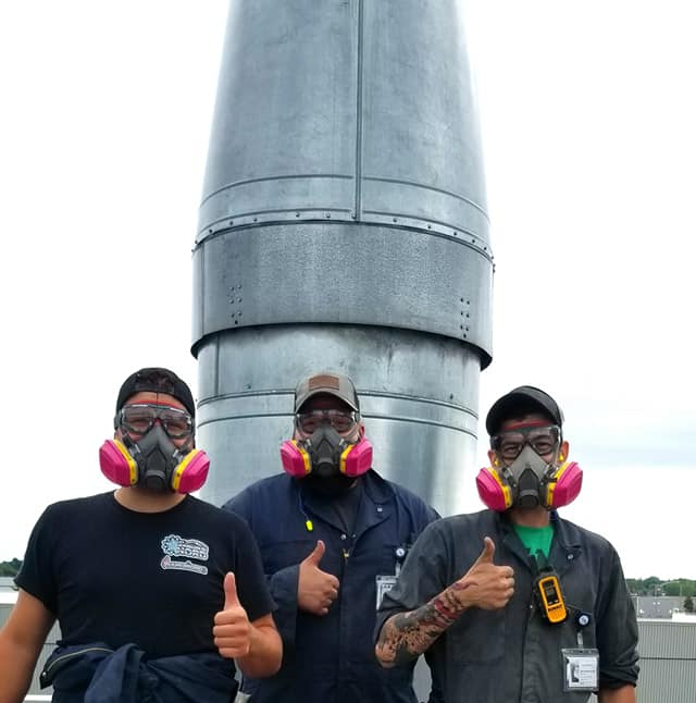 Trois personnes portant des lunettes de sécurité et des masques respiratoires se tiennent devant une grande grille d'aération métallique, levant toutes le pouce.