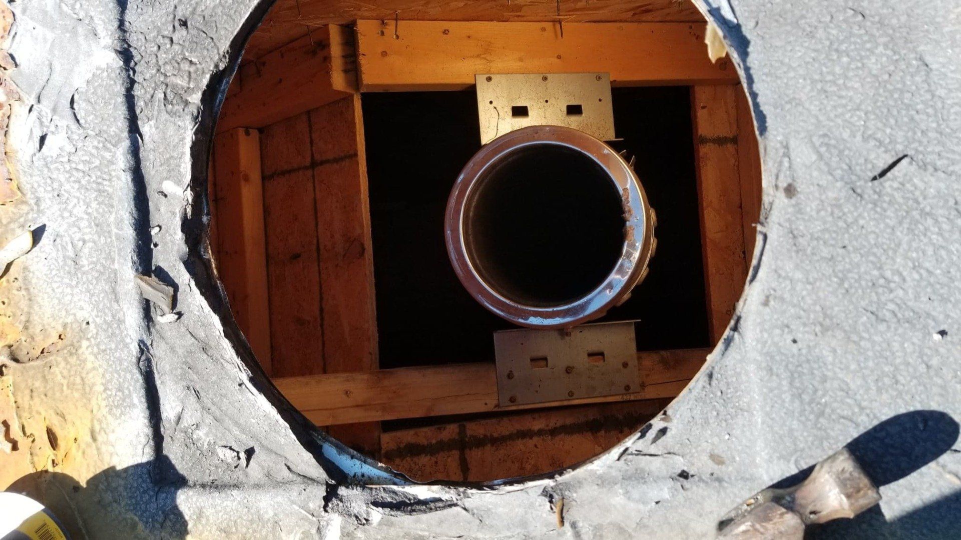 Vue plongeante à travers une ouverture circulaire dans un toit, montrant un conduit de cheminée métallique soutenu par une charpente en bois et des supports métalliques.