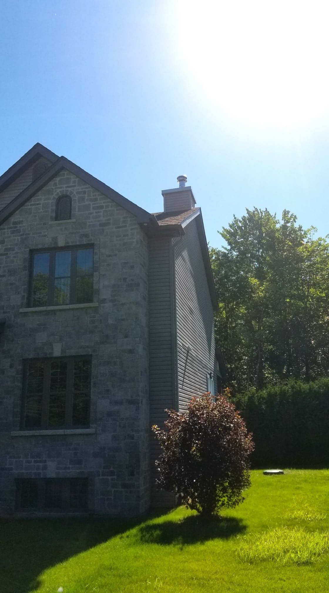 Une maison en pierre, au toit à pignons et au bardage gris, se dresse sur une pelouse verdoyante sous un soleil éclatant, dans un ciel d'un bleu limpide.