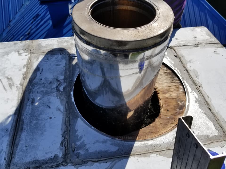 Un conduit de cheminée en acier inoxydable dépasse d'un trou percé dans une toiture en bois, elle-même entourée d'une couronne de cheminée en béton.