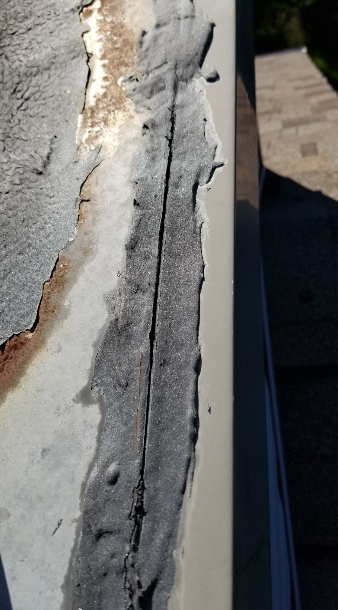 Gros plan sur un joint de solin de toit gris, usé et fissuré, avec un mastic défaillant le long d'un bord métallique.