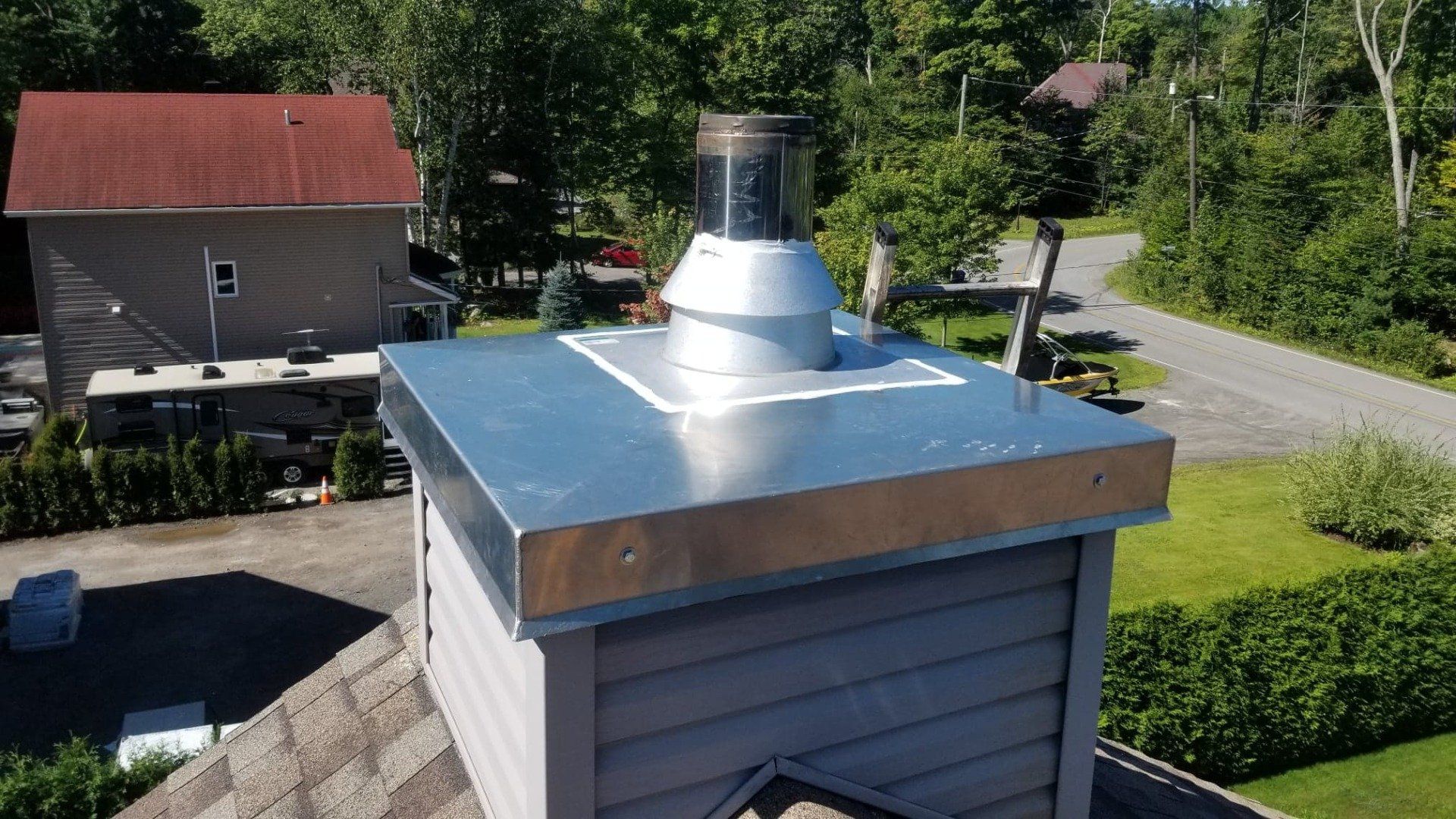 Une cheminée à chapeau métallique sur un toit recouvert de revêtement en vinyle gris, avec une sortie d'air circulaire centrale se détachant sur un fond d'arbres.