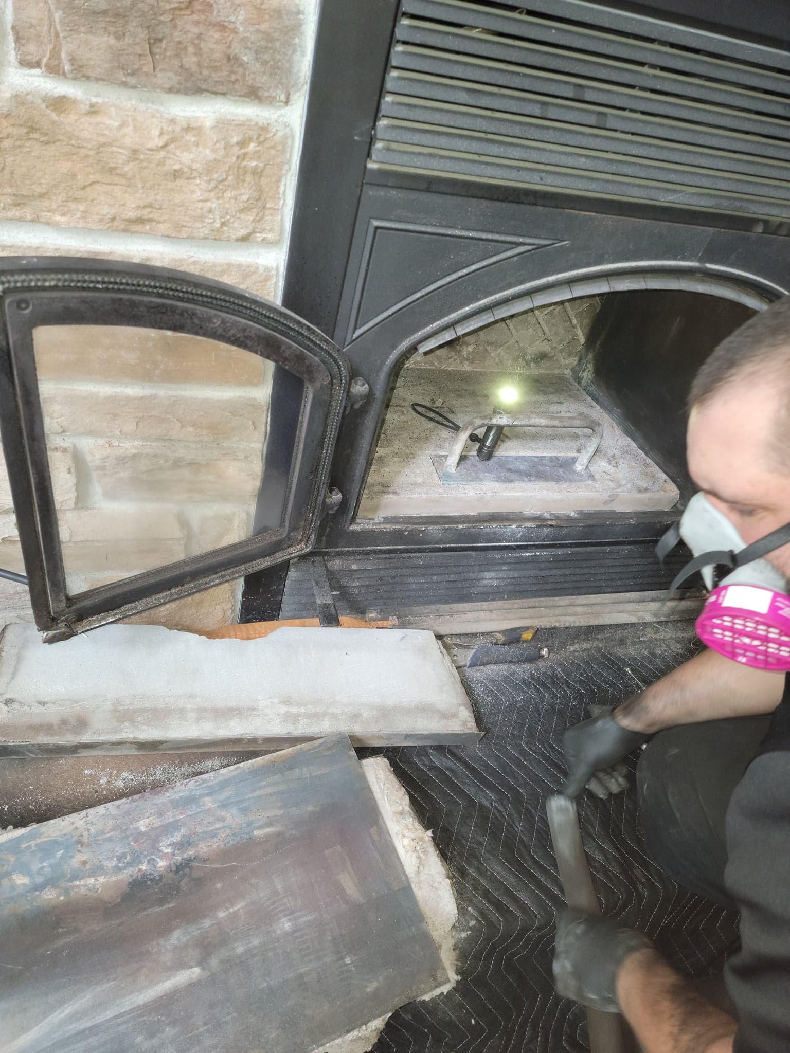 Une personne portant un masque respiratoire nettoie un insert de cheminée à l'aide d'un aspirateur, une dalle de foyer étant placée au sol.