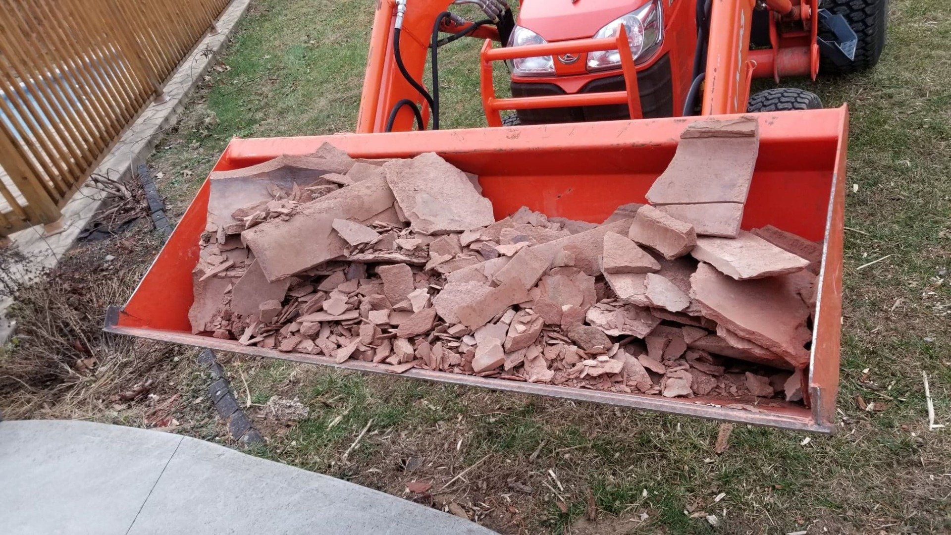 Un godet de chargeur de tracteur orange vif rempli de morceaux de grès brun rougeâtre brisés, posé à l'extérieur sur l'herbe.