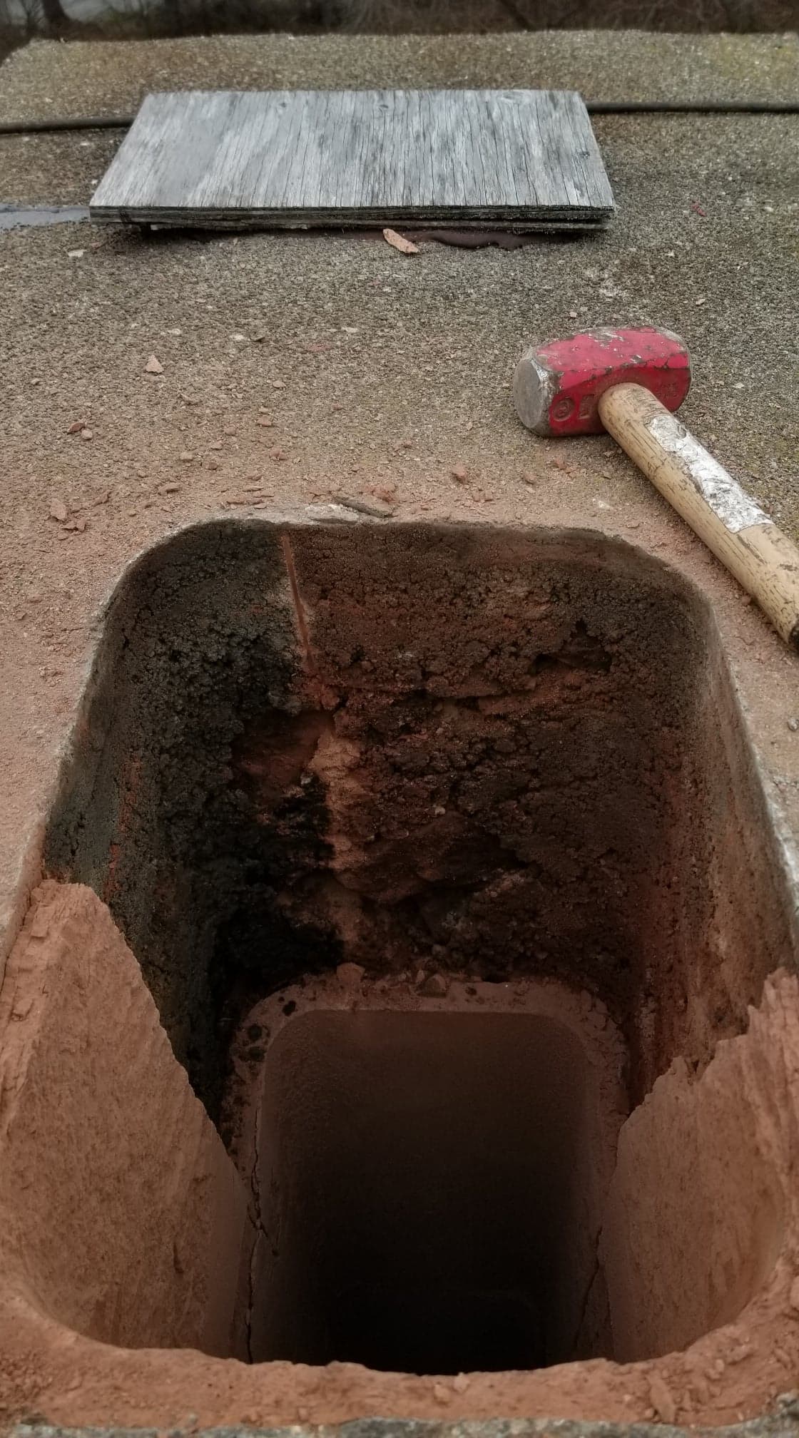 Vue de dessus d'un conduit de cheminée montrant une importante accumulation de suie, avec un maillet rouge et une tuile plate à proximité sur le toit.