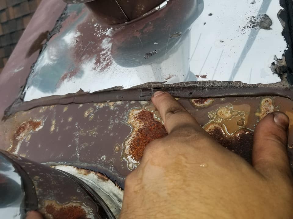 Une personne montre du doigt un défaut d'étanchéité dans le joint d'étanchéité le long d'une solin de cheminée métallique rouillée sur un toit.