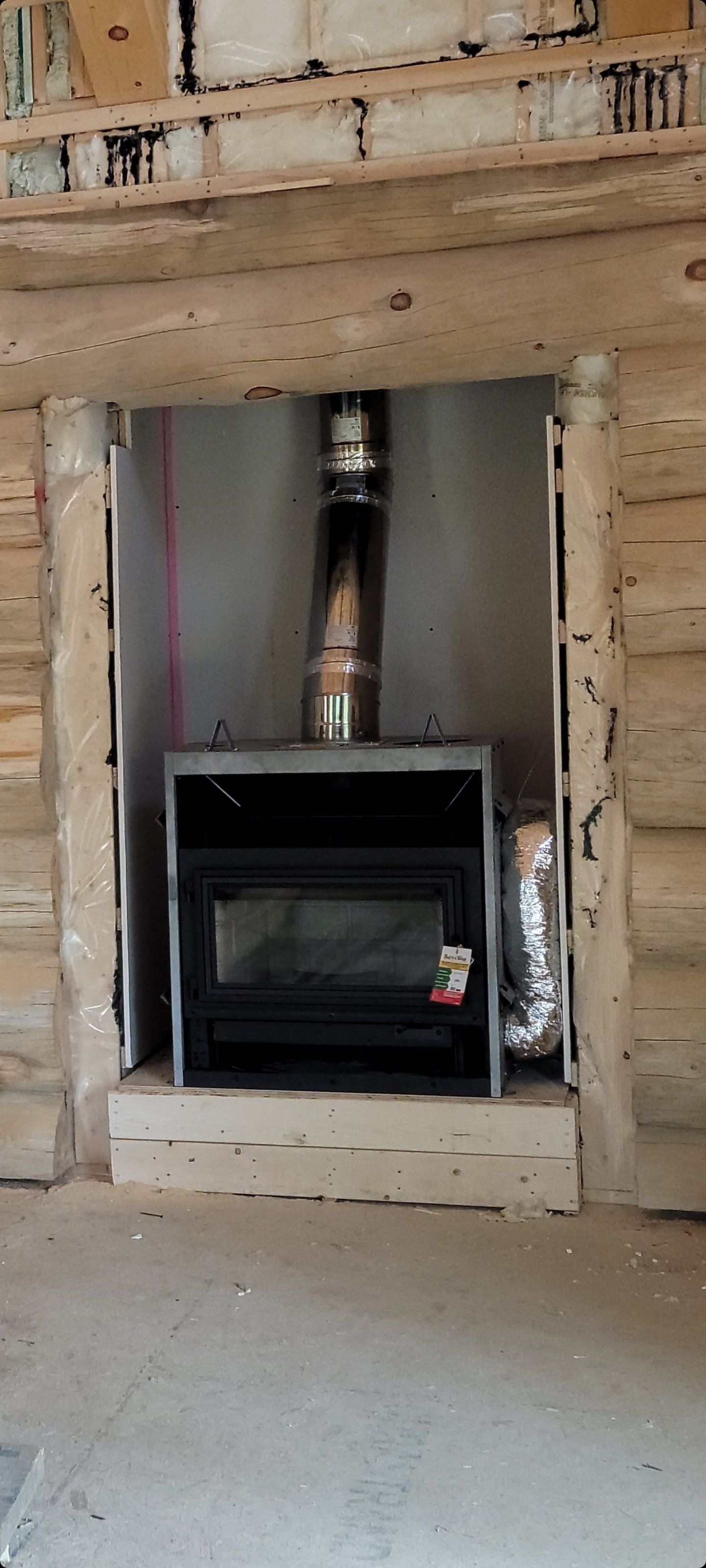 Un insert de cheminée à bois noir installé dans une alcôve non finie à l'intérieur d'un mur en rondins, avec une cheminée métallique apparente.