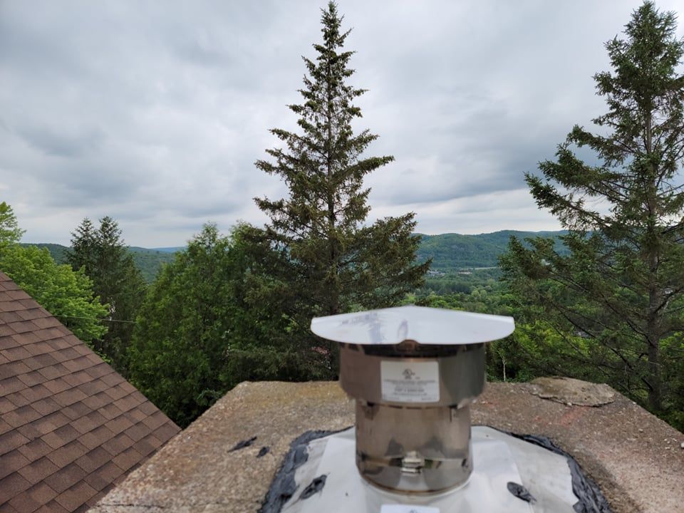 Un chapeau de cheminée en acier inoxydable repose sur un socle en béton, surplombant une vallée boisée sous un ciel nuageux.