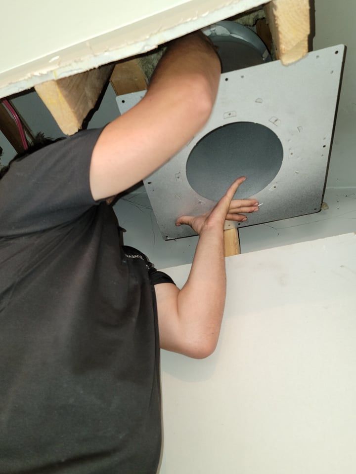 Une personne installe un support de plafond métallique pour un ventilateur de ventilation entre les solives de plafond en bois.