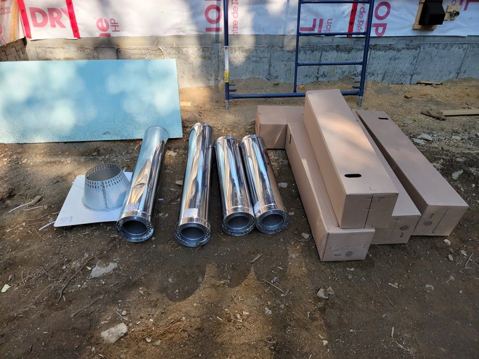 Chantier de construction avec des sections de tuyaux de cheminée métalliques, une base de solin et des cartons d'expédition posés sur un sol en terre battue.