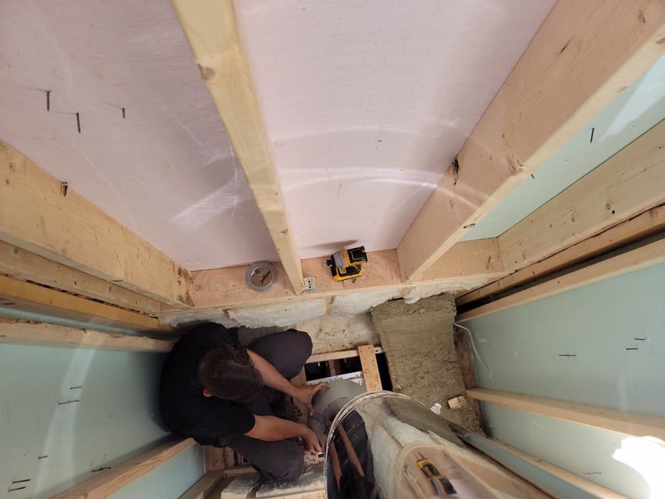 Une personne travaille à l'installation d'un tuyau métallique dans un espace de construction étroit, délimité par des poutres en bois et des panneaux isolants.