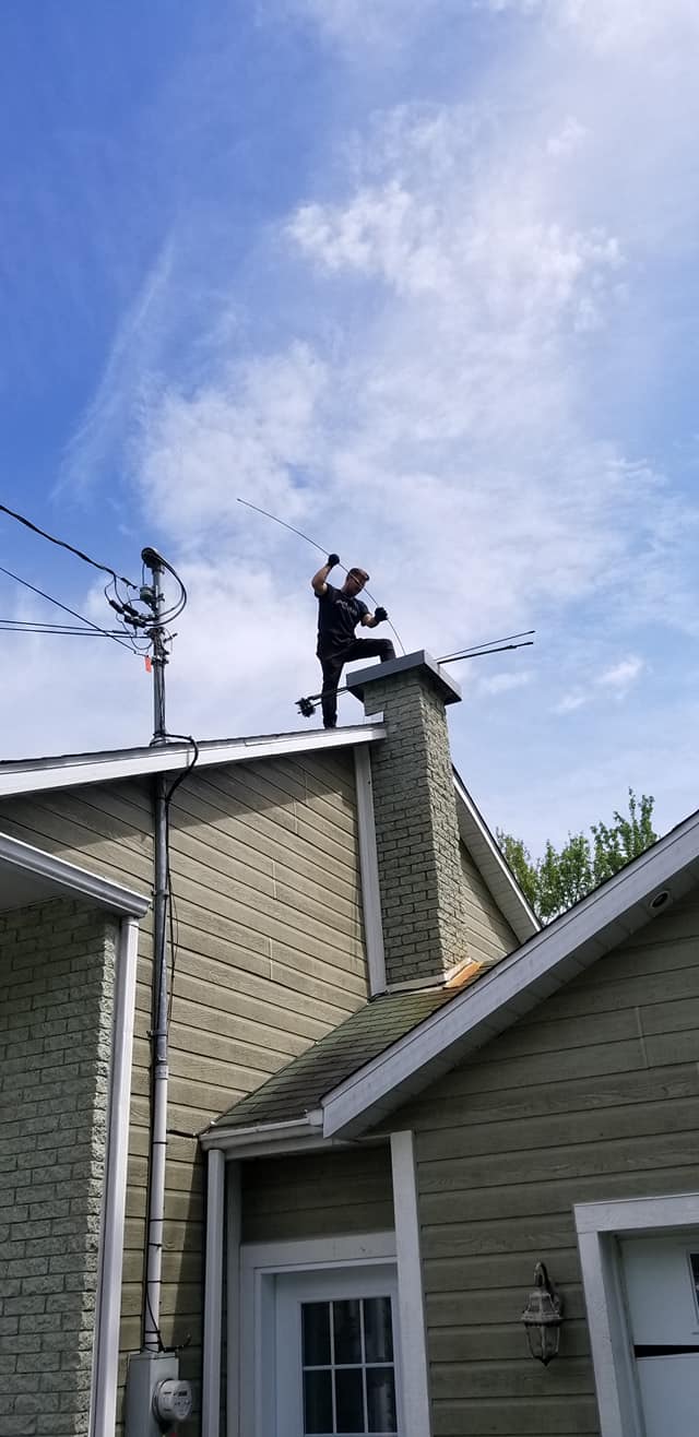 Une personne se tient sur le toit d'une maison, près d'une cheminée, et utilise un long outil pour dégager les débris sur fond de ciel bleu nuageux.