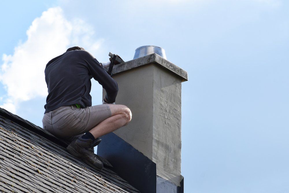 Une personne vêtue d'une chemise grise et d'un short est accroupie sur le toit d'une maison pour travailler sur une cheminée, sur fond de ciel bleu.