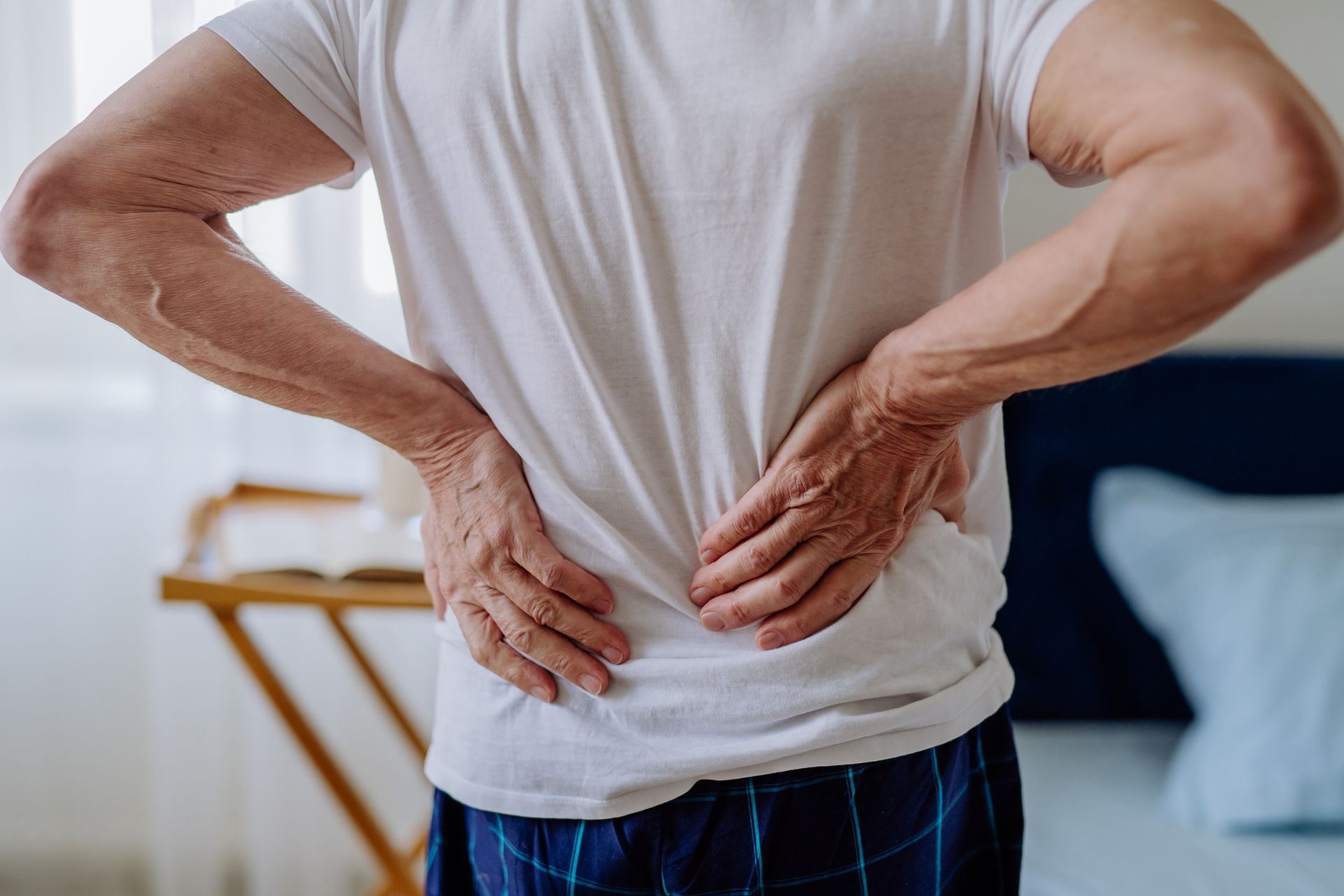 Man in white shirt holding his lower back, presumably in pain. Indoors, near a bed.