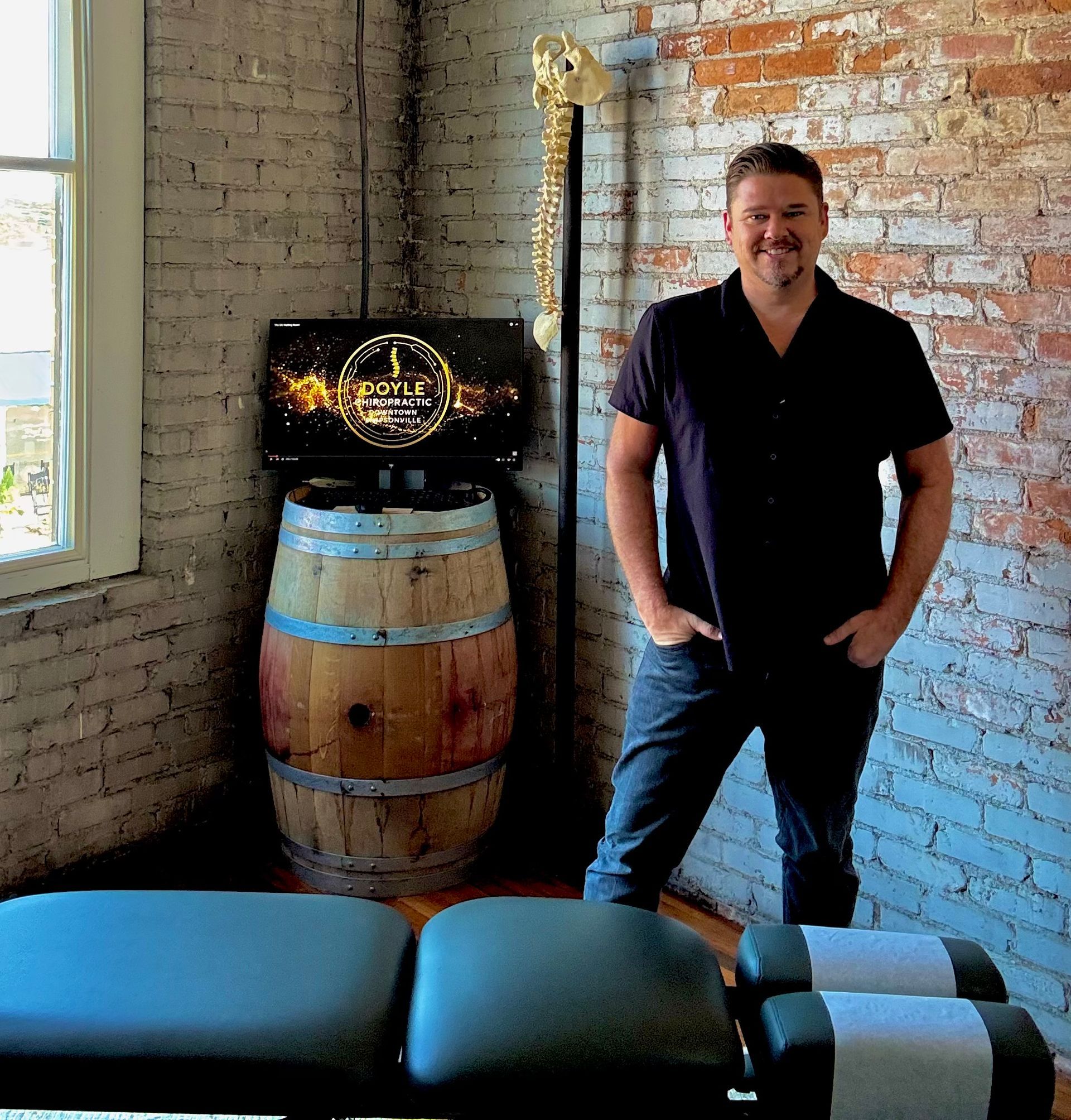 Chiropractor in a black shirt, standing in front of a wine barrel with a spinal column model. Exposed brick wall.