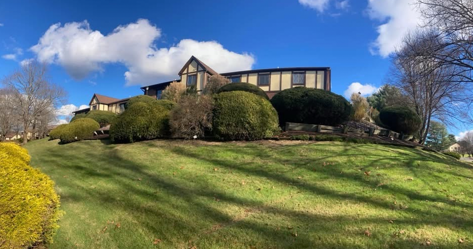 Tudor style house on a hill. Brown roof, beige siding, and green lawn under a blue sky with clouds.