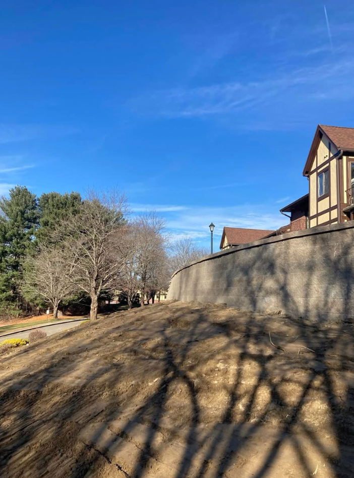 Blue sky over a brown-roofed building, a long wall, bare trees, and a brown dirt hillside.