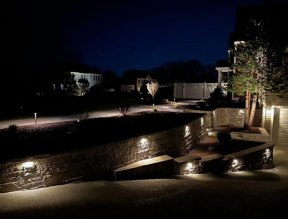 Nighttime view of a landscaped yard with illuminated stone walls, pathways, and landscaping.