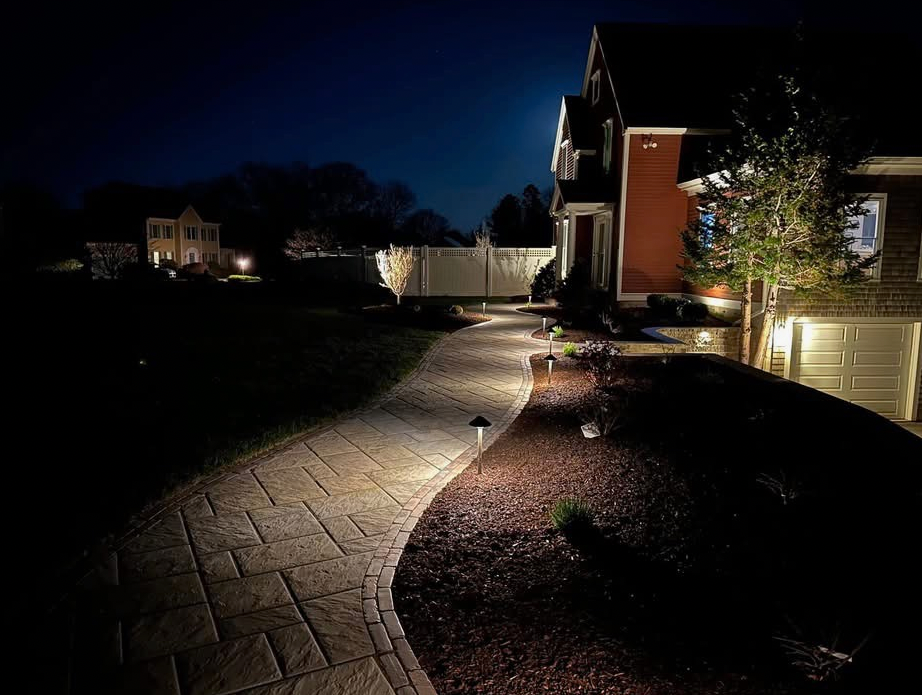 Brick pathway illuminated by lights leading to a house with a red exterior at night.