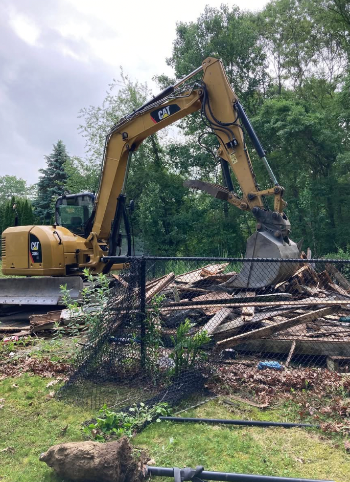 Yellow excavator demolishing debris near a fence in a yard. Green trees and cloudy sky in background.