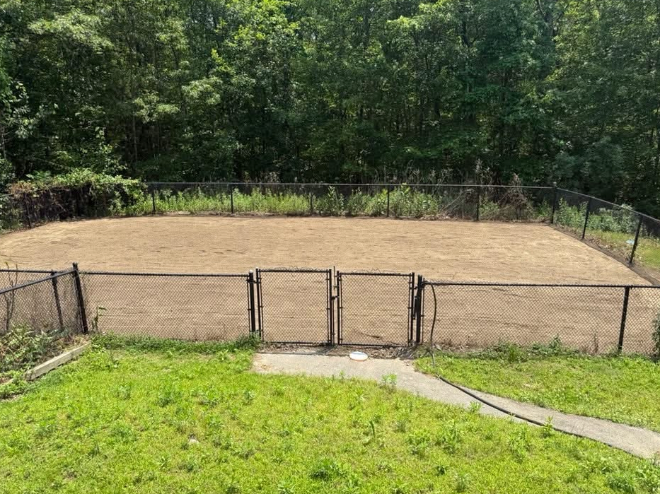 A fenced-in area of dirt with a gate, backed by trees and green grass.