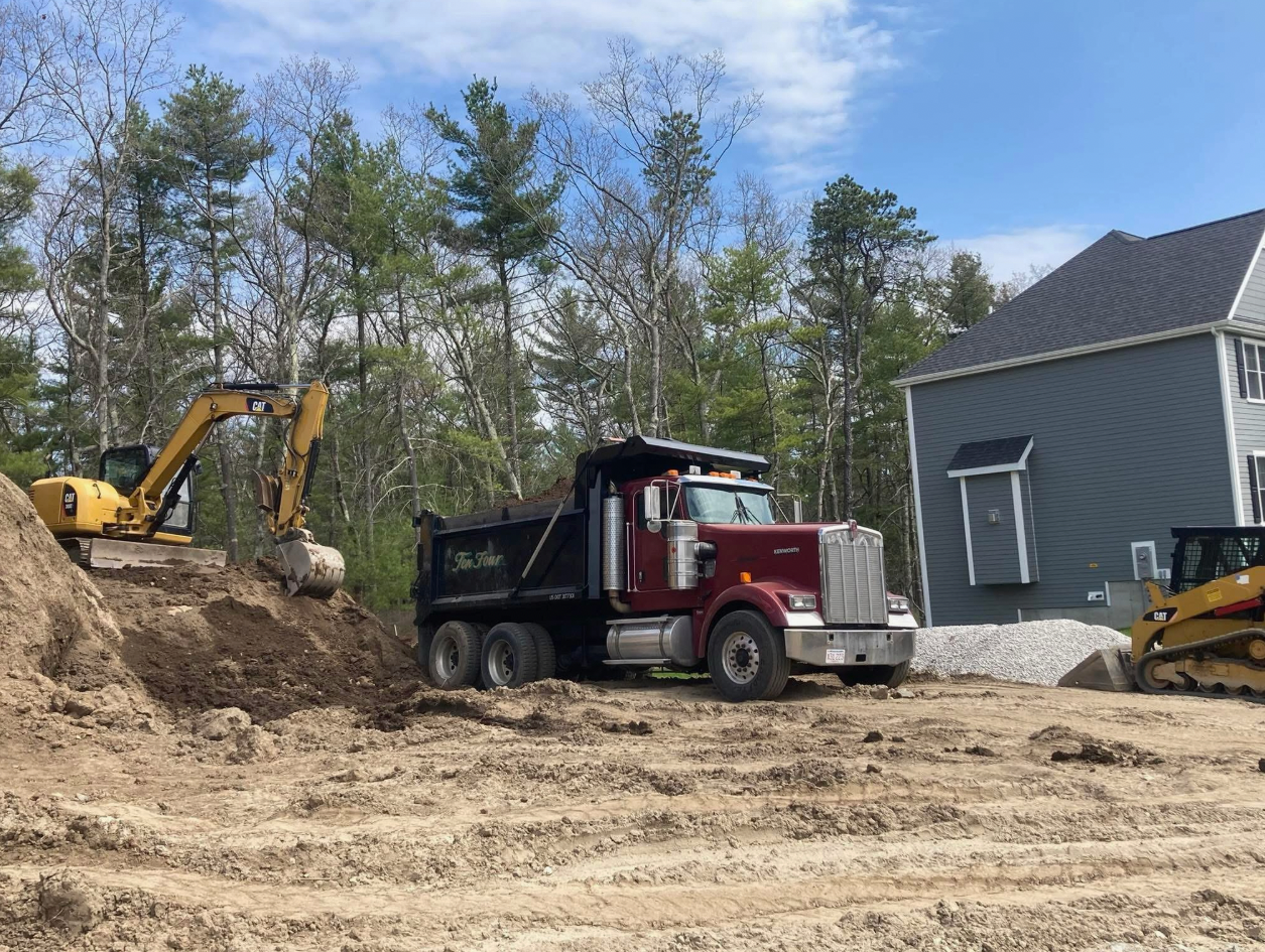 Excavator loading dirt into a maroon dump truck next to a house under construction.
