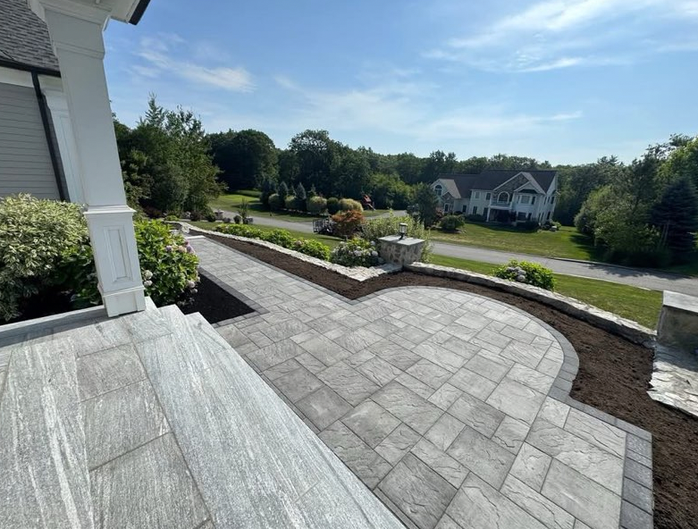 Stone patio with curved retaining walls, landscaping, and houses in the distance under a blue sky.