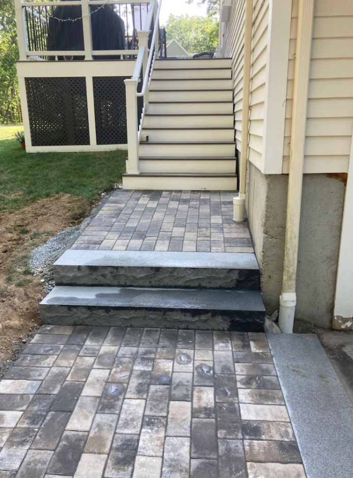 Stone pavers leading to stairs under a deck with light-colored siding and stairs.