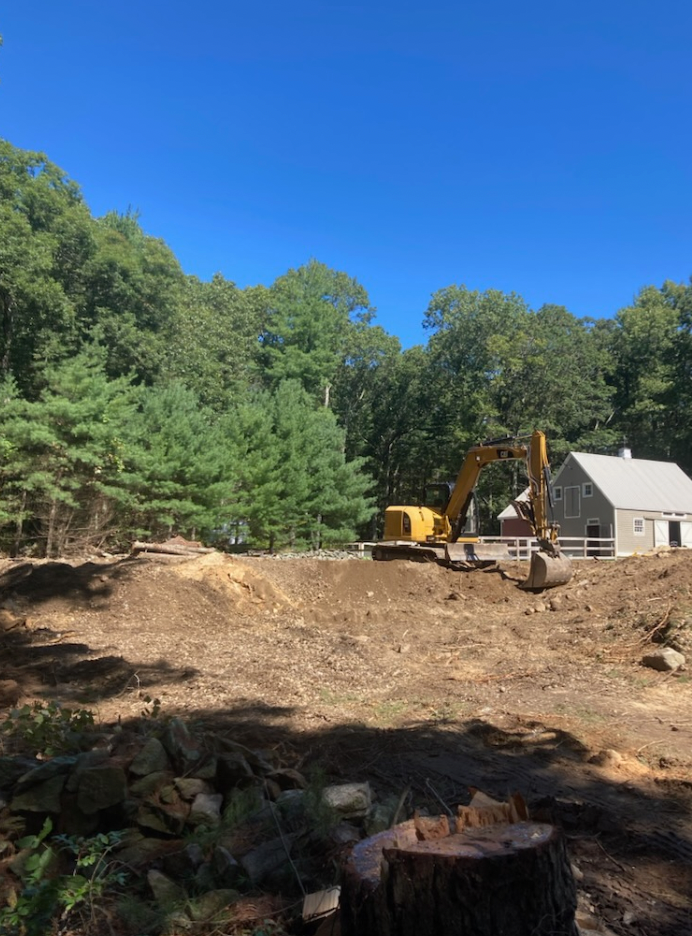 A yellow excavator on a dirt construction site clearing land near a house, surrounded by trees under a blue sky.