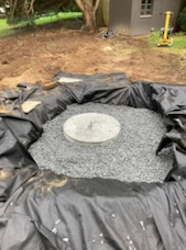Black liner and gravel surround a concrete circle in a dug-out area, outdoors.