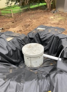A concrete cylinder with holes sits in a black liner within an excavated area, next to a white pipe.