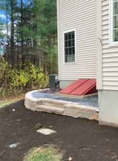 Exterior view of a house with a red basement door and a stone retaining wall with steps.