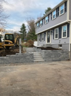Gray block retaining wall with steps leading to a gray house with blue shutters, a yellow excavator nearby.
