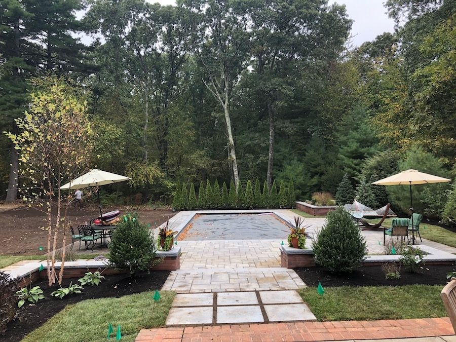 Patio with pool, landscaping, trees, seating, and umbrellas. Green grass, brick and stone.