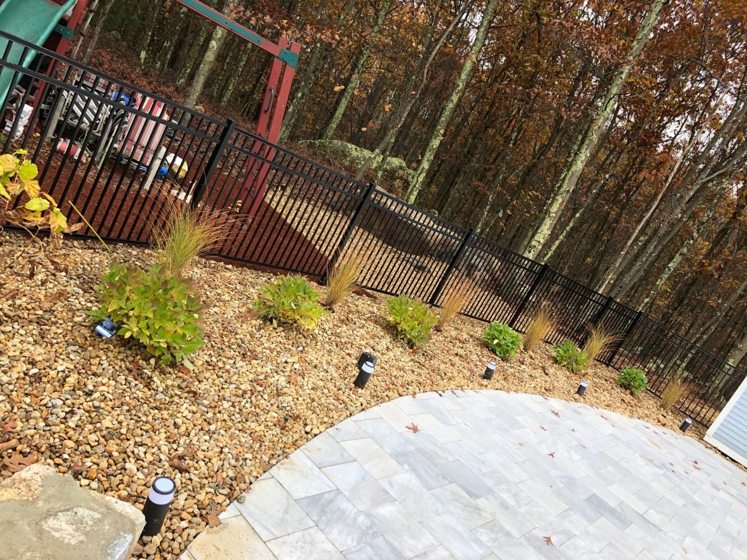 Stone patio with ground cover and plants, bordered by a black fence and trees.
