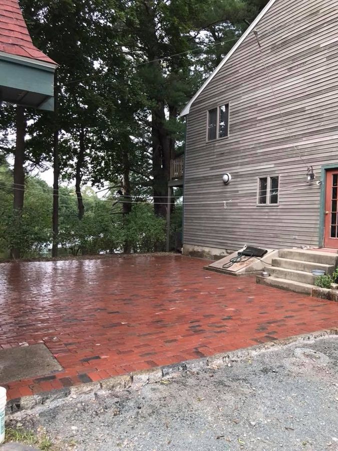 Brick patio wet from rain next to a wooden house with steps and a view of trees and water.